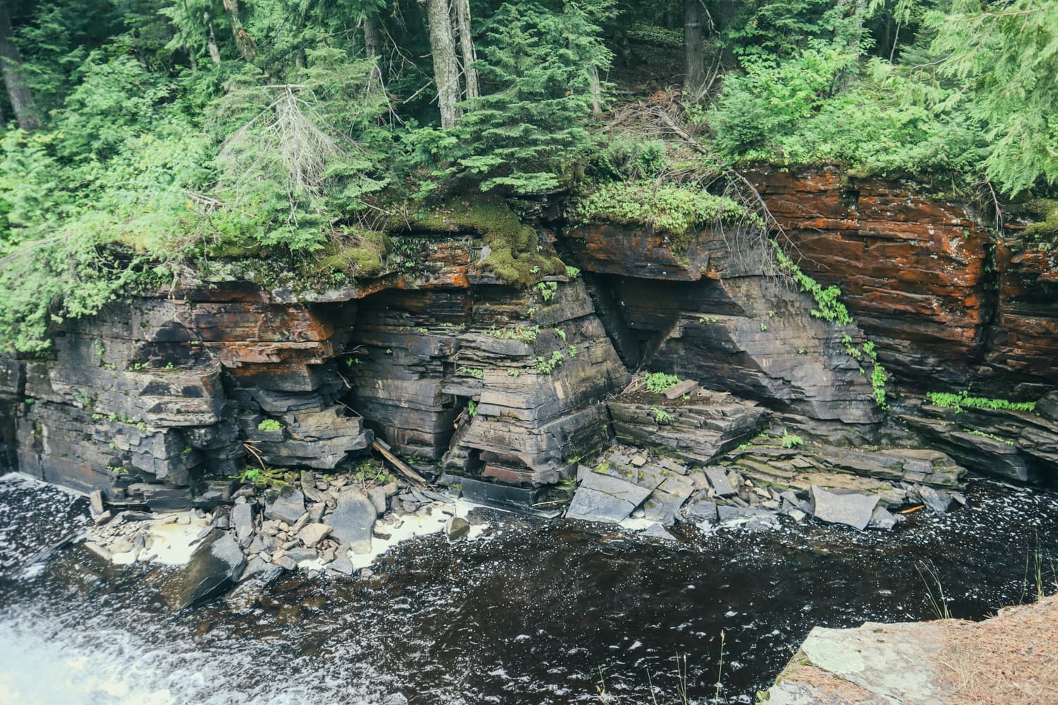 Layered rust-colored rock walls of the Sturgeon River gorge rising above the water at Canyon Falls