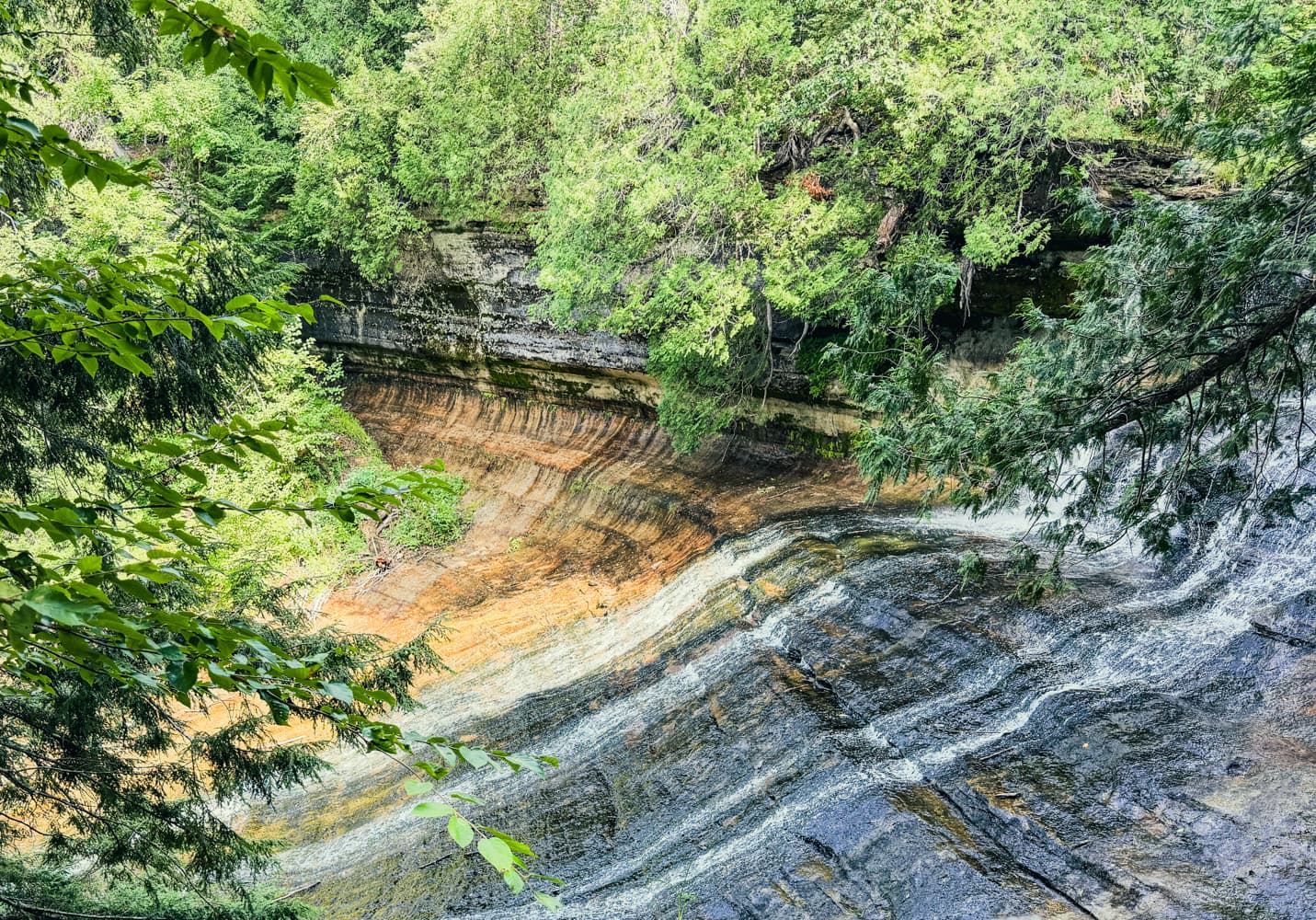 Wide view of the Laughing Whitefish Falls gorge showing orange and gray layered rock beneath the forest canopy