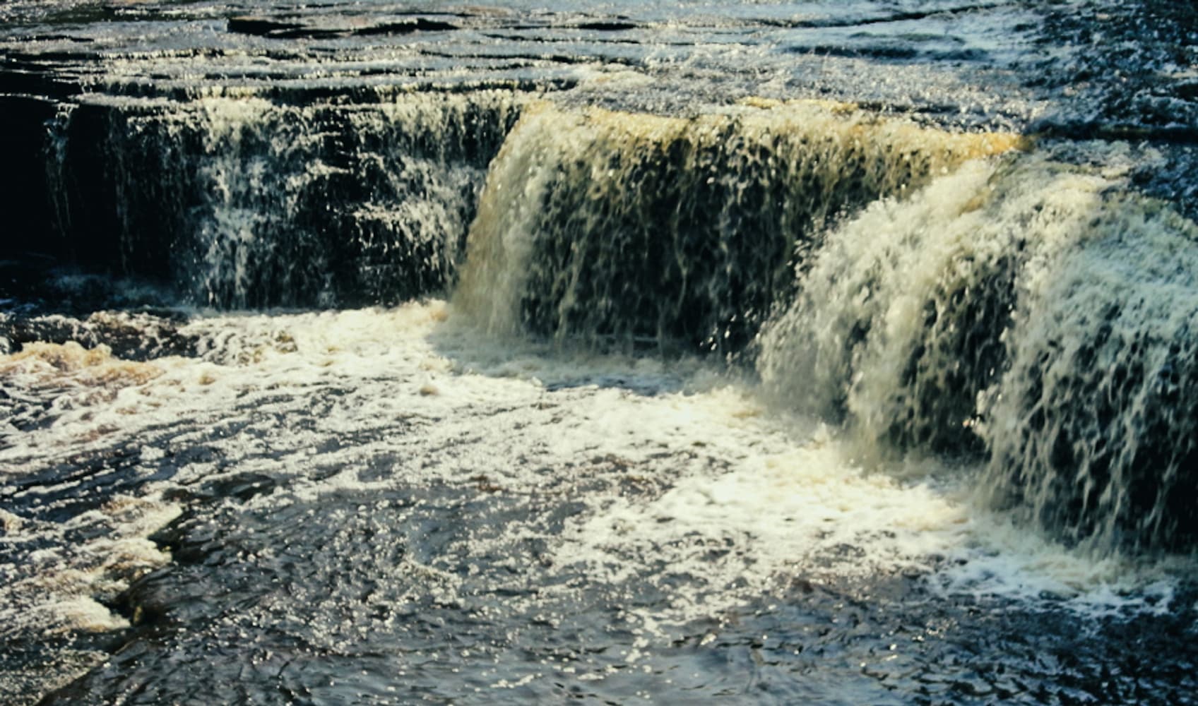 Close view of amber Tahquamenon River water cascading over a dark rock ledge