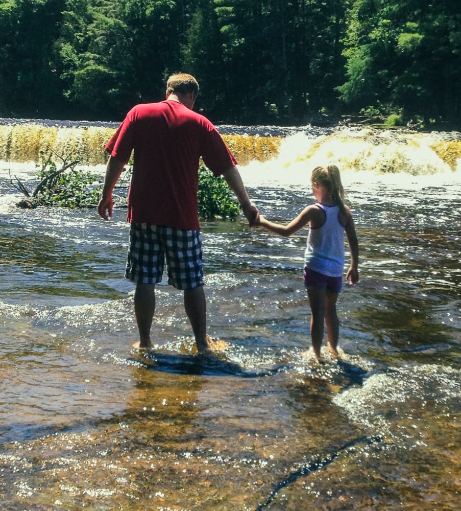 A father and daughter wading hand in hand toward the cascades at Tahquamenon Lower Falls