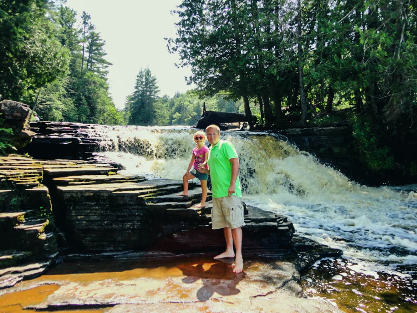 A father and daughter standing on the layered rock ledges beside Tahquamenon Lower Falls