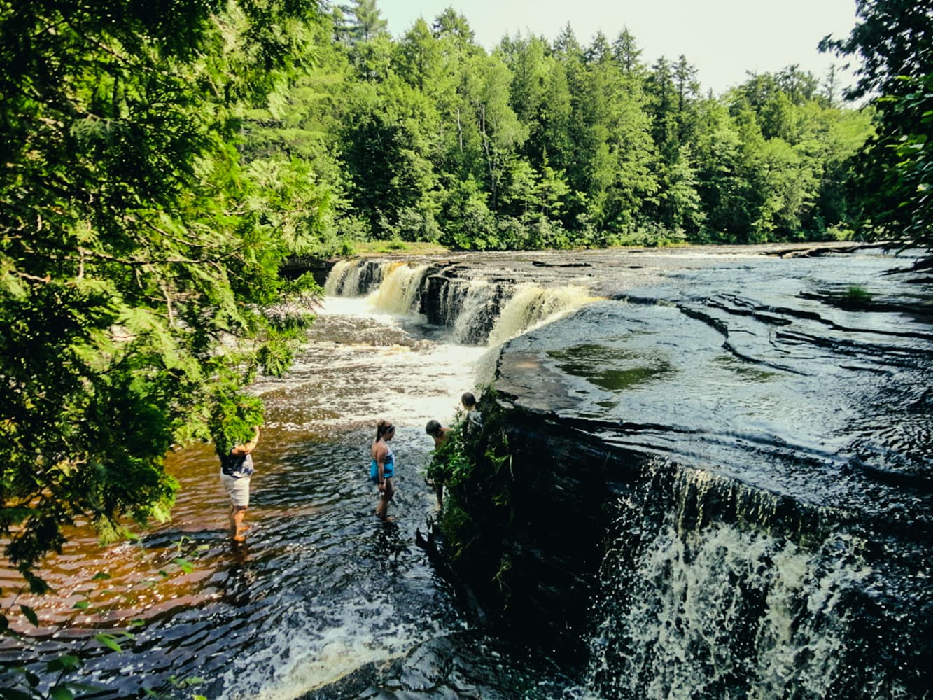 Tahquamenon Lower Falls cascading through a forested channel as visitors wade in the river below